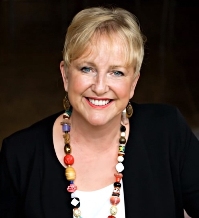 Portrait of a smiling woman with blonde hair, wearing a black top and a colorful beaded necklace.