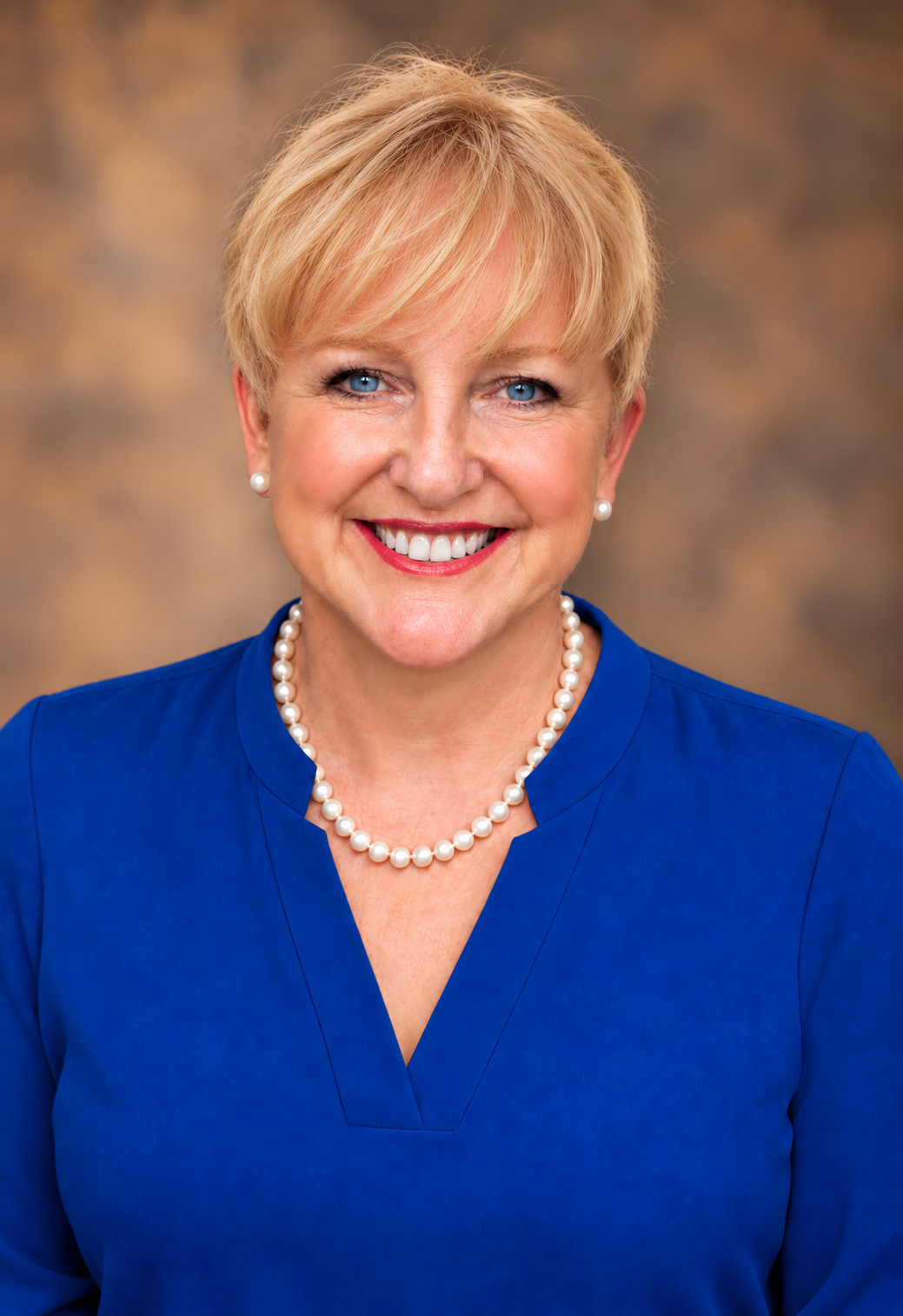 Smiling blonde woman in a royal blue blouse with a pearl necklace and matching earrings, posing for a headshot.